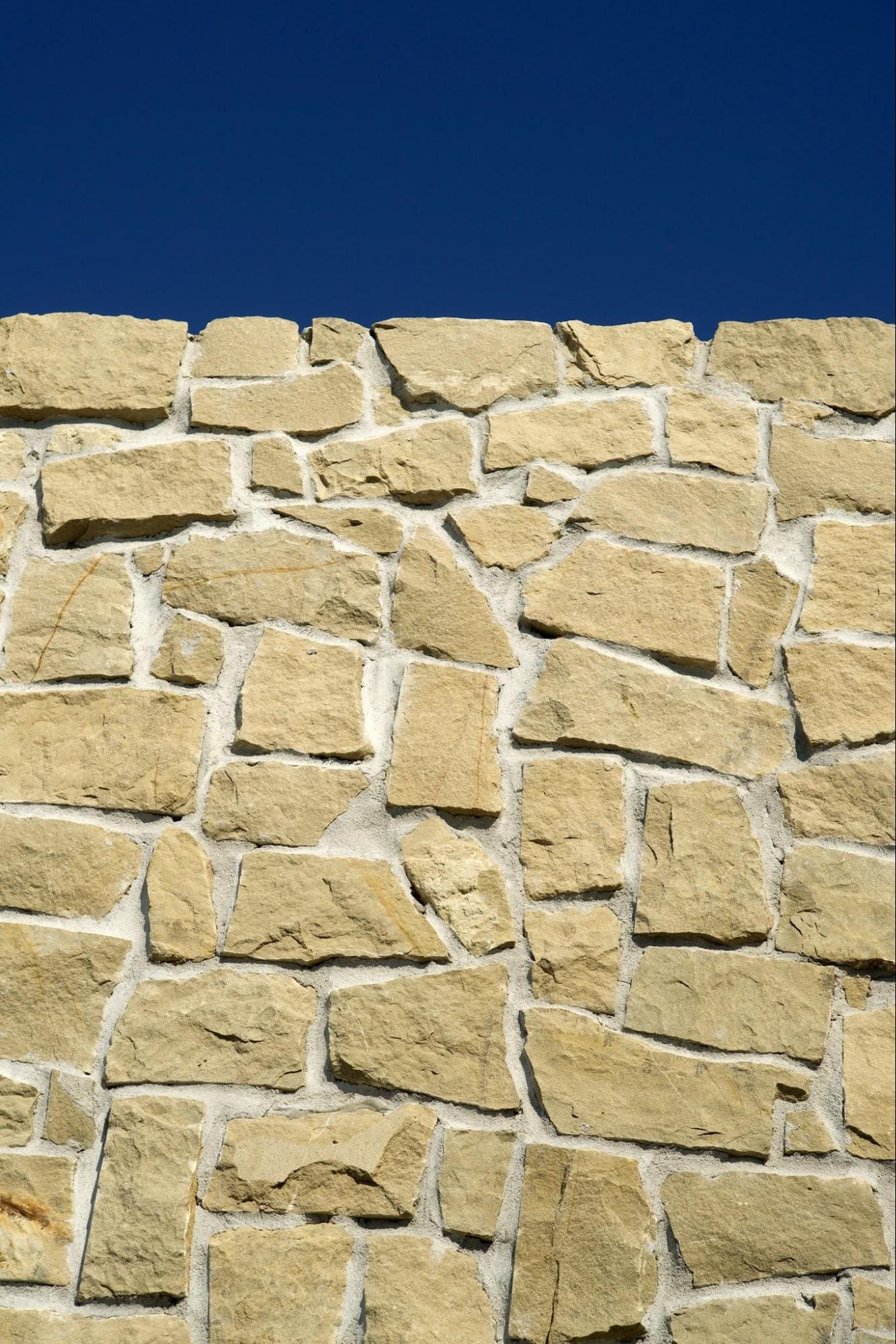 A durable natural stone wall built against a bright blue sky. Because natural stone varies in size and shape, each piece must be carefully selected, positioned, and fitted by a mason to create a consistent appearance.