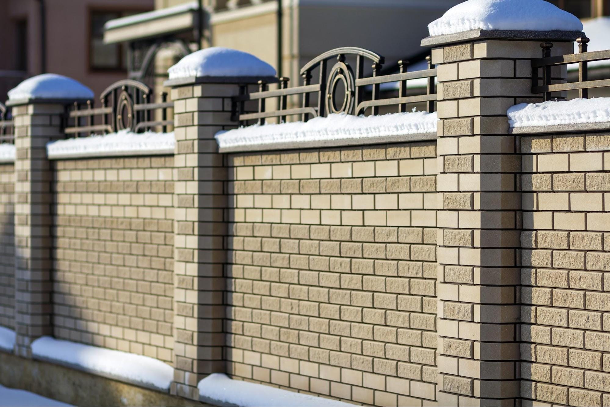 A solid masonry wall featuring prominent stone columns and decorative metal fencing covered in a light dusting of snow. Stone columns frequently serve as focal points for fence lines and pair exceptionally well with ornamental steel fencing.